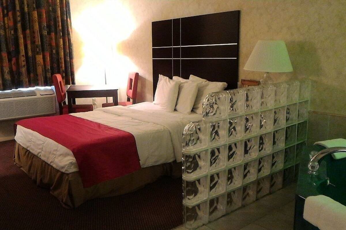 A hotel room with a bed, a bedside table, a lamp, a glass cabinet with decorative items, and a window with curtains.