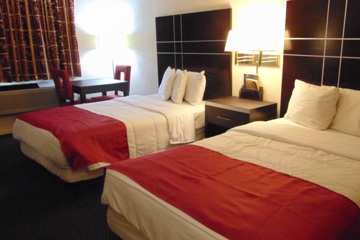 A hotel room with two beds, each with white bedding and red accents, a small desk with a chair, and a nightstand with a lamp.