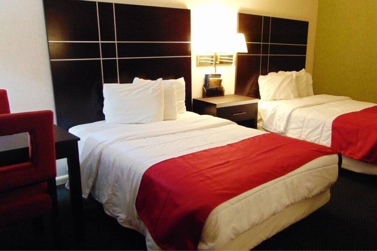 A hotel room with two beds, a red chair, a nightstand with a lamp, and a wall-mounted clock.