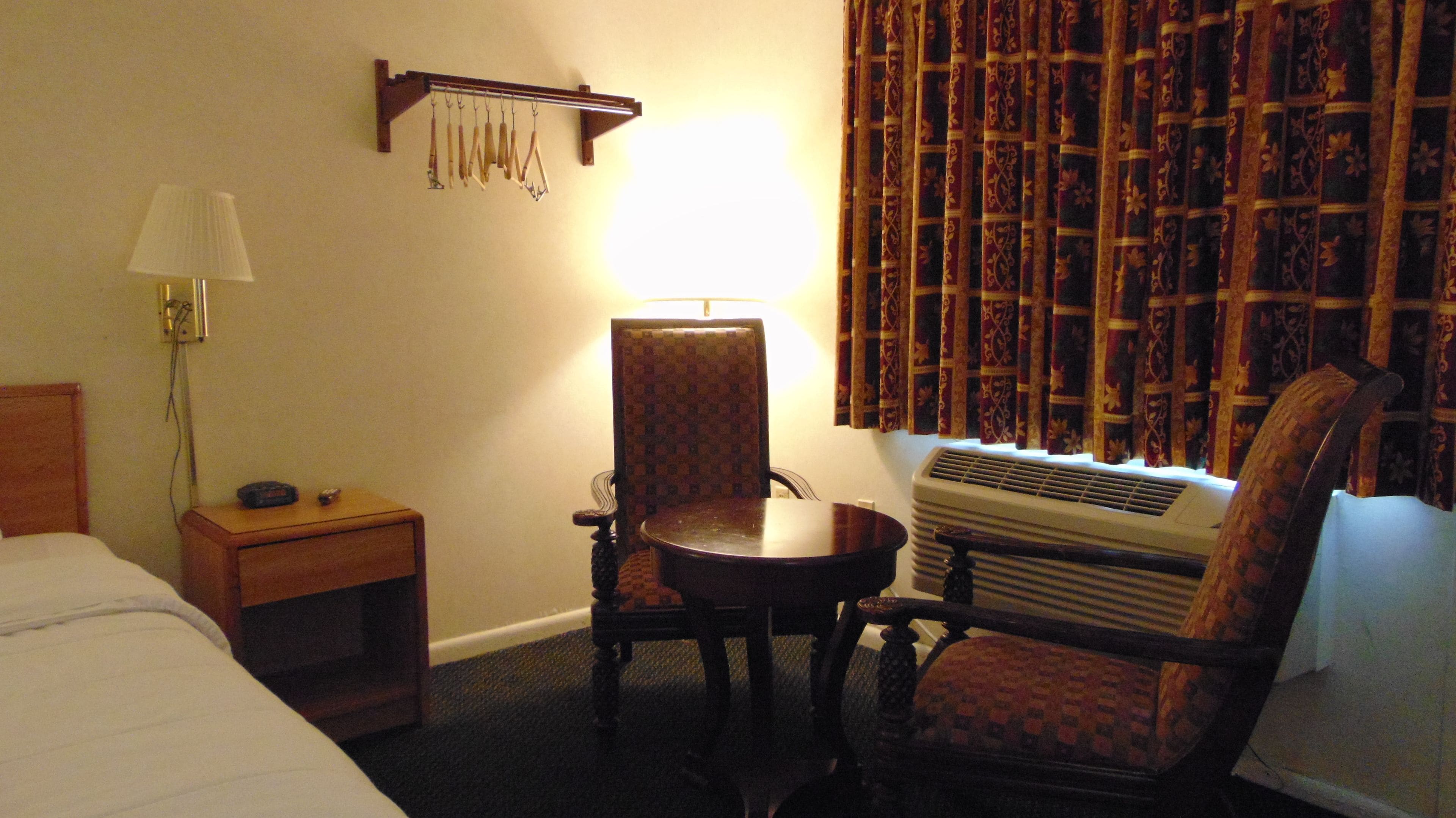 Habitación de hotel con cama, mesita de noche, escritorio, silla, mesa, lámpara, aire acondicionado y ventana con cortinas estampadas.
