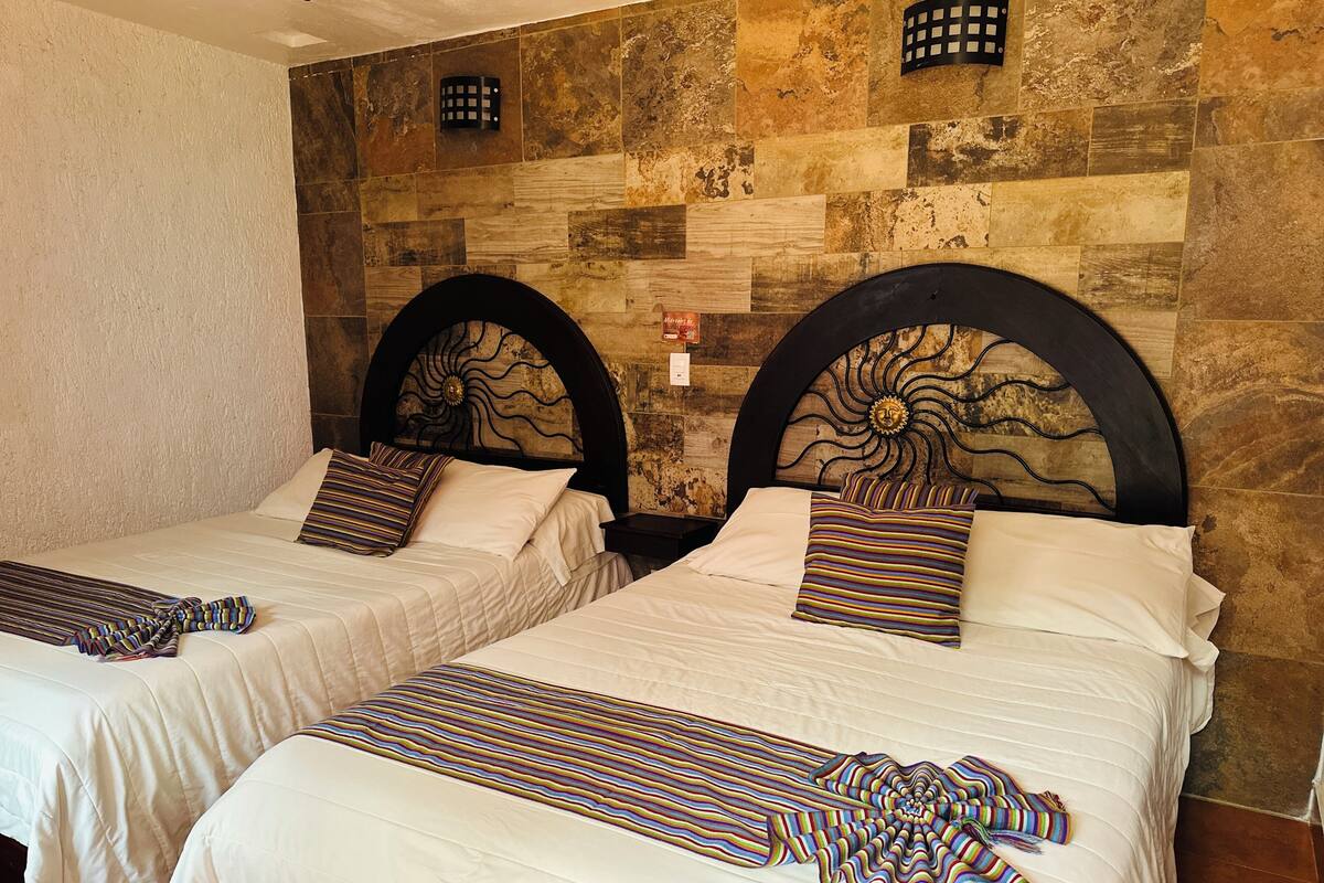 Two twin beds with striped bedspreads and matching pillows, set against a stone wall with a sun design on the headboard.