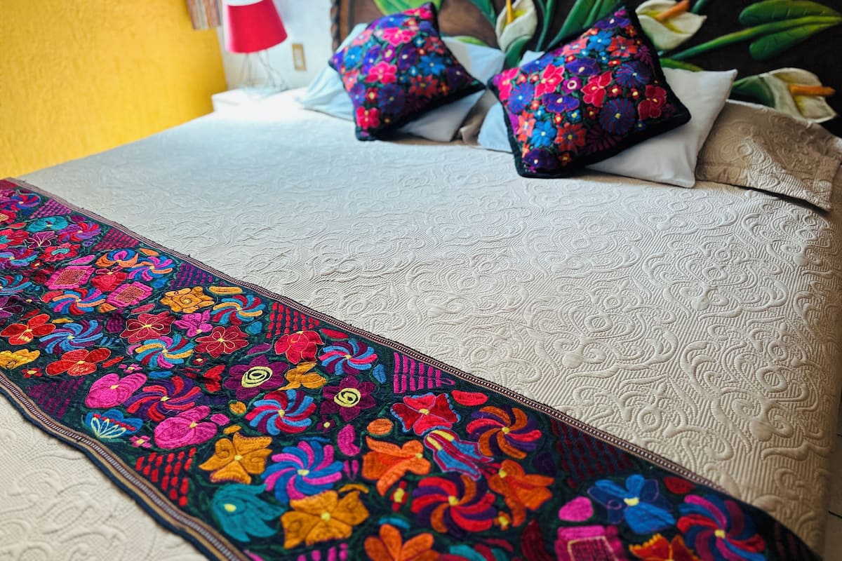 A bed with a colorful embroidered runner, two pillows with a floral pattern, and a red lamp in the background.