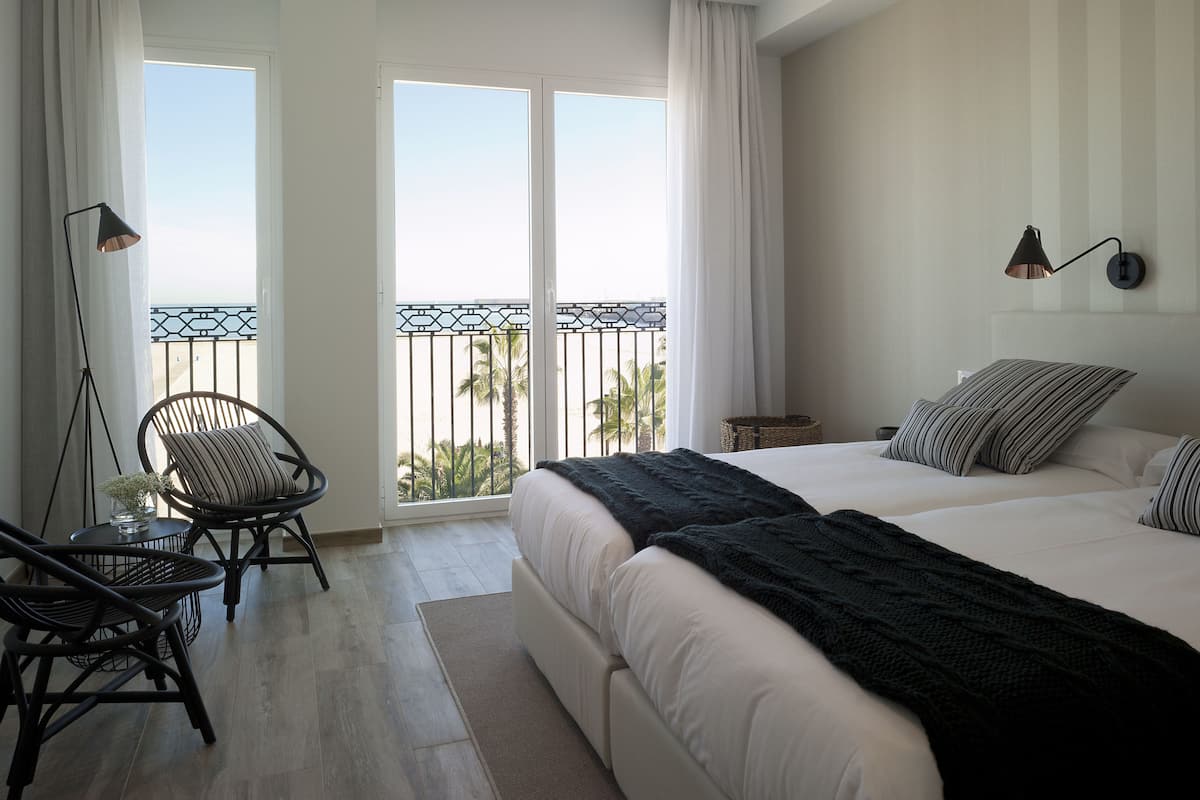 A bedroom with a large bed, a chair, a floor lamp, and a view of the outdoors through a balcony.
