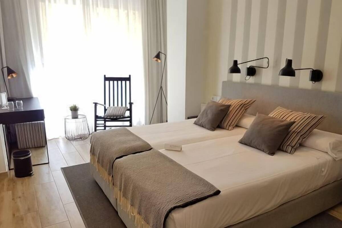 A double bed with a headboard, bedside tables, a chair, and a desk with a lamp.