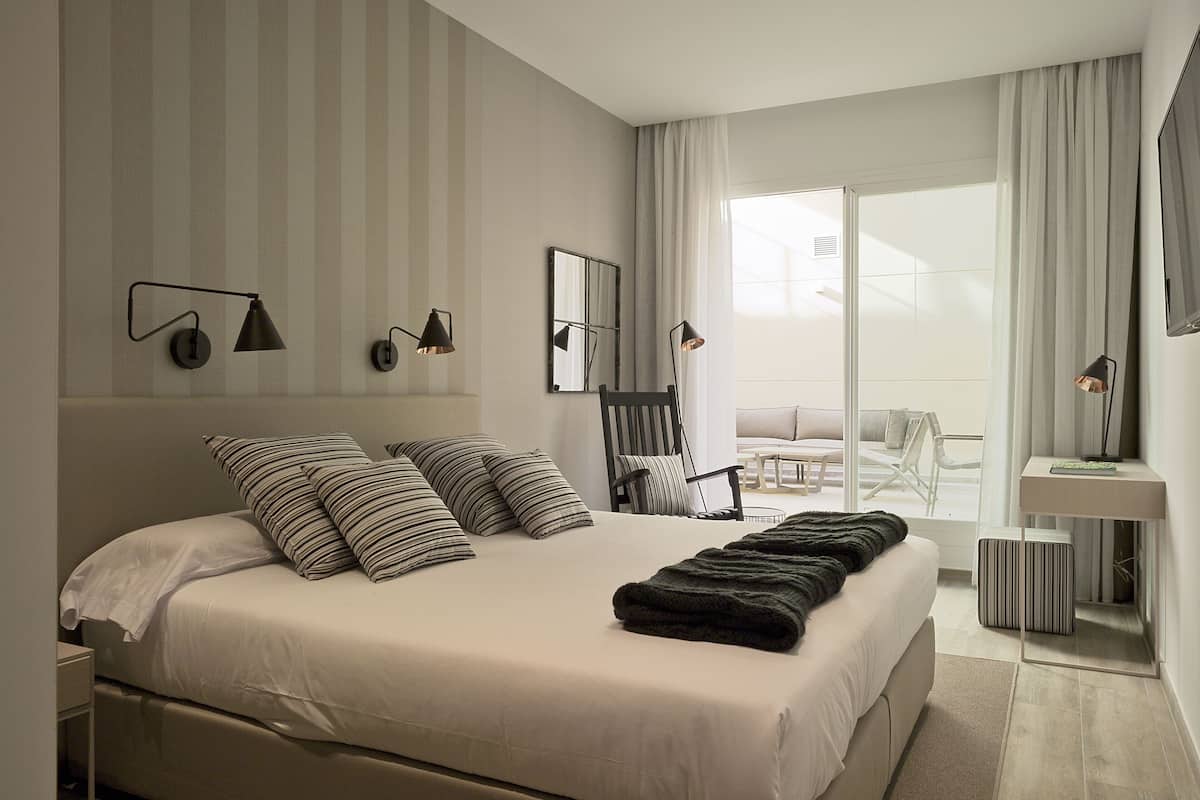 A modern hotel room with a large bed, a chair, a small table, and a view of the outdoors.