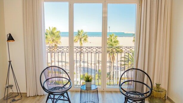 Una habitación con un gran ventanal con vistas a la playa, dos sillas negras, una pequeña mesa con una planta y una lámpara de pie.