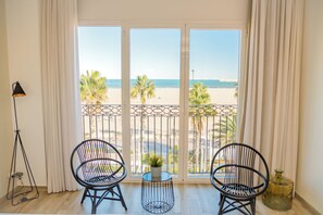 Una habitación con un gran ventanal con vistas a la playa, dos sillas negras, una pequeña mesa con una planta y una lámpara de pie.
