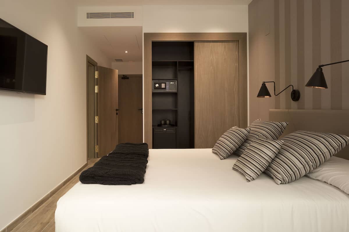 A modern bedroom with a large bed, a black throw blanket, and striped pillows. There is a wardrobe, a small kitchen area, and a TV mounted on the wall.