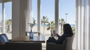 Una habitación con un gran ventanal con vistas a la playa, una mesa con un florero, y una persona leyendo un libro.