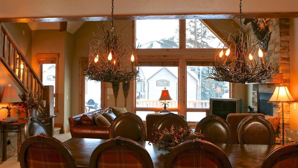 Un coin repas avec une table et des chaises en bois, un lustre et une vue sur une maison, visible à travers de grandes fenêtres.
