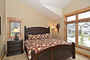 A bedroom with a wooden bed, a nightstand with a lamp, a dresser, and a window with a view of houses. - Lakota Antlers 200 5 Bedroom Holiday Home by Winter Park Lodging Company (Winter Park)