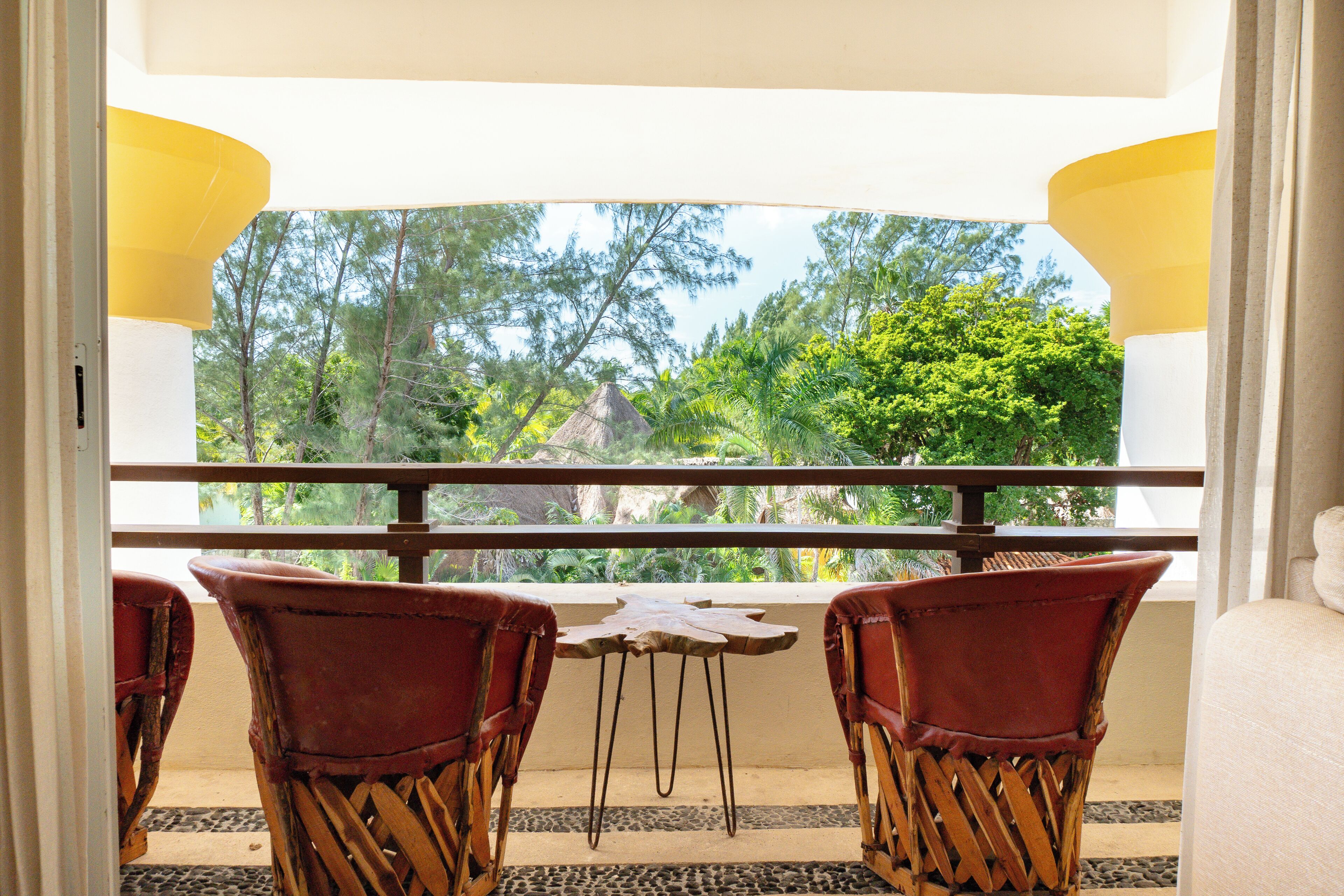 Un balcon avec deux fauteuils en osier et une petite table, donnant sur un paysage verdoyant et luxuriant, avec des arbres et une construction au toit de chaume.