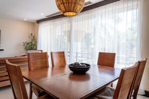 Un comedor con una mesa de madera, cuatro sillas, un cuenco de fruta y un ventanal con cortinas ligeras.