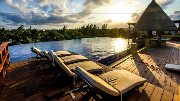 A wooden deck with lounge chairs and a thatched-roof structure by a pool.