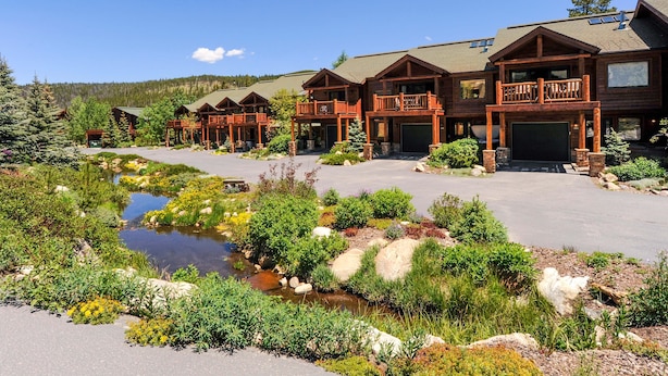 A lodge with multiple wooden buildings, a small stream, and a mountainous backdrop. - Slopeside 202 3 Bedroom Holiday Home by Winter Park Lodging Company (Winter Park)