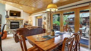 A cozy living room with a fireplace, wooden ceiling, and a large window overlooking a deck. - Slopeside 202 3 Bedroom Holiday Home by Winter Park Lodging Company (Winter Park)
