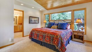A bedroom with a large bed, wooden ceiling, two bedside tables with lamps, a window with a view of trees, and a door leading to a bathroom. - Slopeside 202 3 Bedroom Holiday Home by Winter Park Lodging Company (Winter Park)