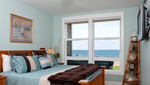 Ein Schlafzimmer mit großem Fenster mit Blick auf das Meer, einem Bett mit vielen Kissen, einem Holz-Kopfteil und einem Regal mit dekorativen Gegenständen.
