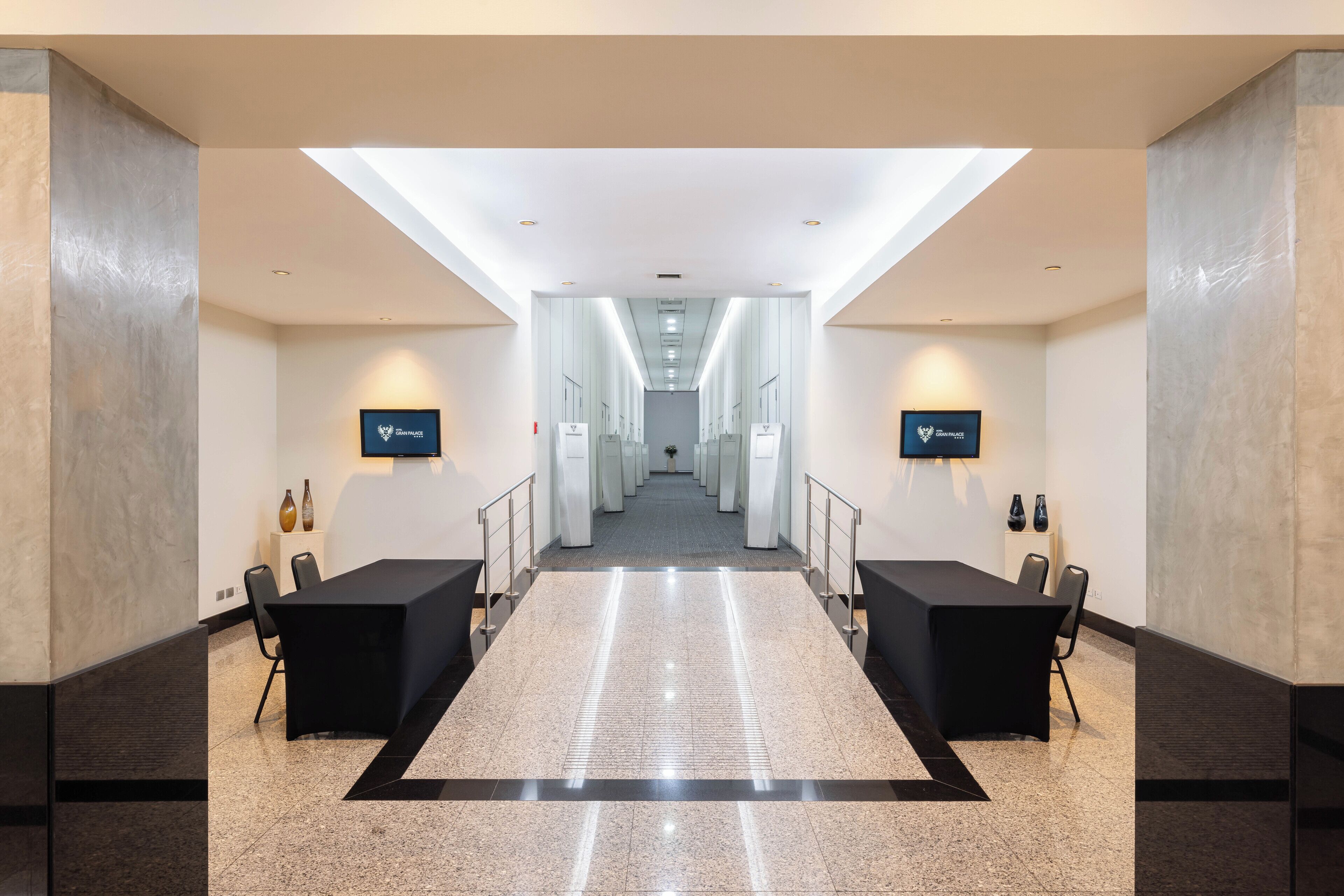A hotel hallway with a central corridor, flanked by two reception desks with chairs, and a ceiling with recessed lighting.