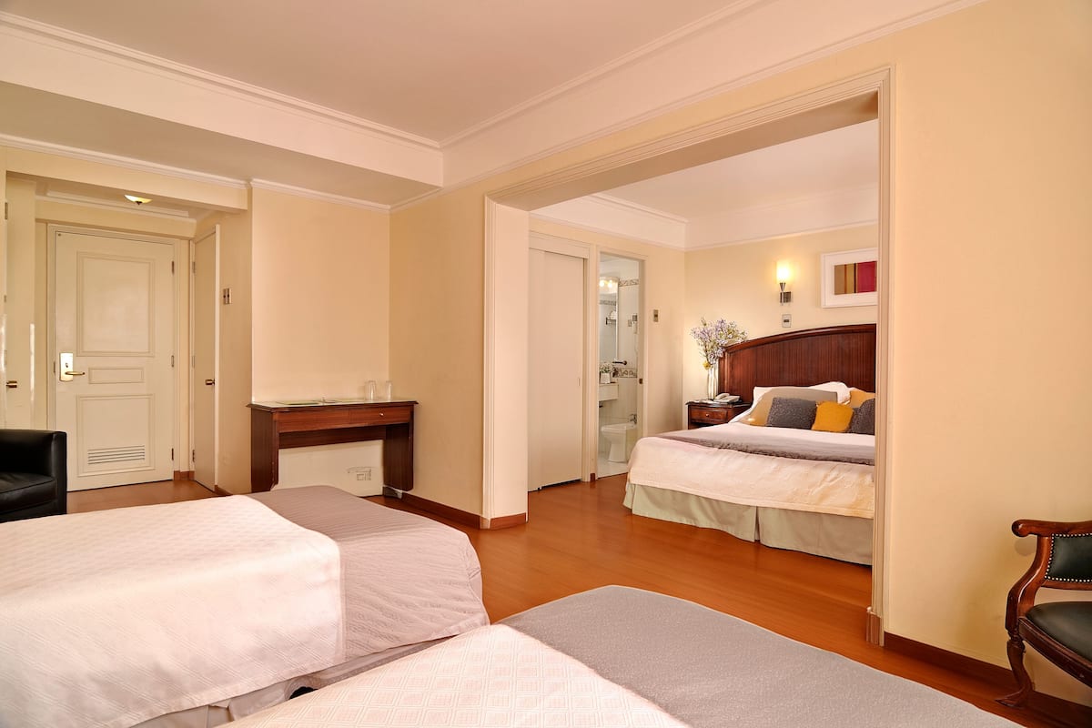 Habitación de hotel con dos camas, un escritorio, una silla y un baño visible a través de una puerta abierta.
