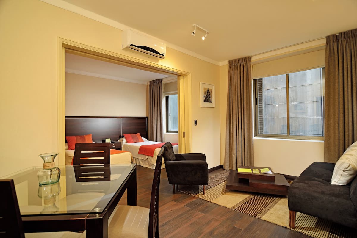 Habitación de hotel con cama, sofá, mesa de comedor y una silla. Cuenta con ventana con cortinas, un cuadro en la pared y un equipo de aire acondicionado.