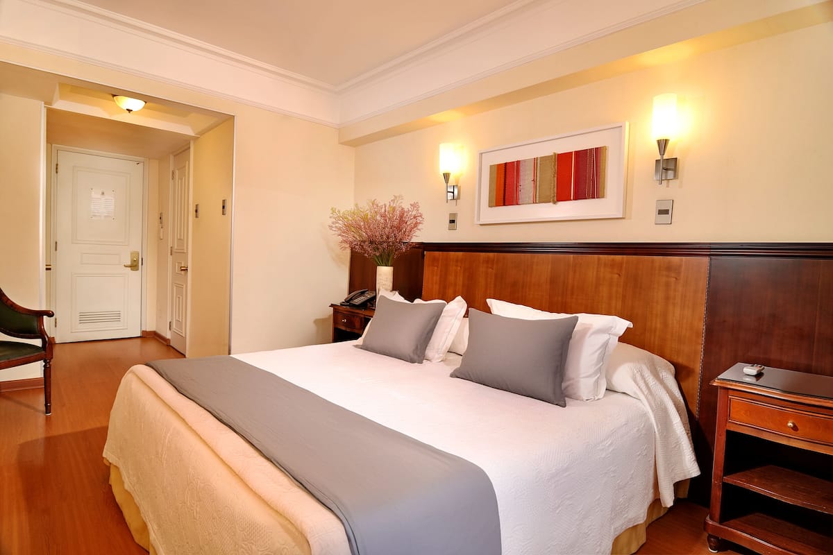 Habitación de hotel con una cama grande, un cabecero de madera, una mesita de noche, una obra de arte enmarcada y un jarrón con flores.