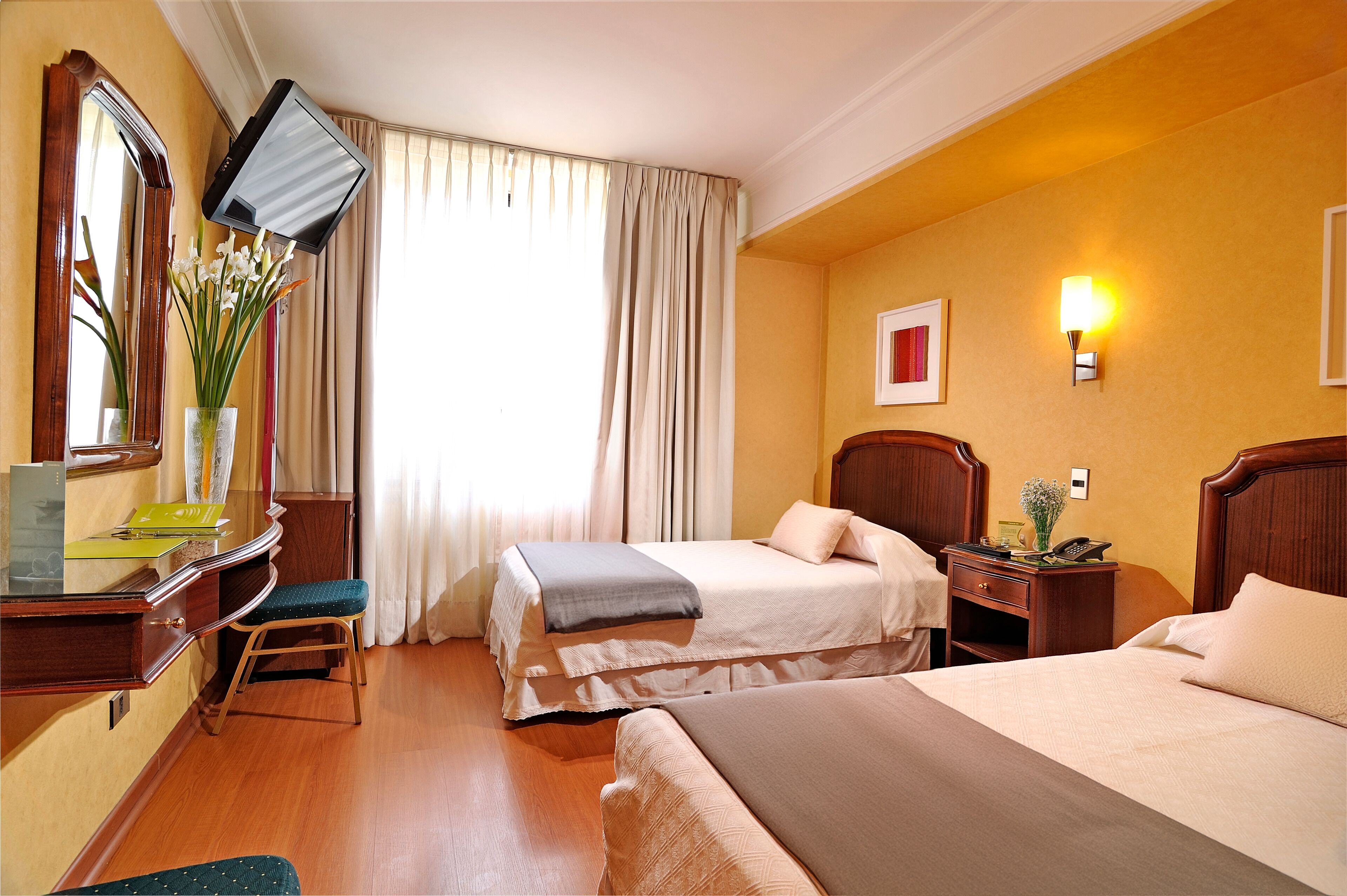 A hotel room with two beds, a desk with a chair, a television mounted on the wall, and a window with curtains.