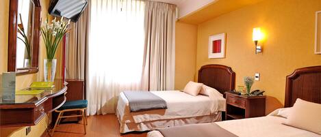 A hotel room with two beds, a desk with a chair, a television mounted on the wall, and a window with curtains.