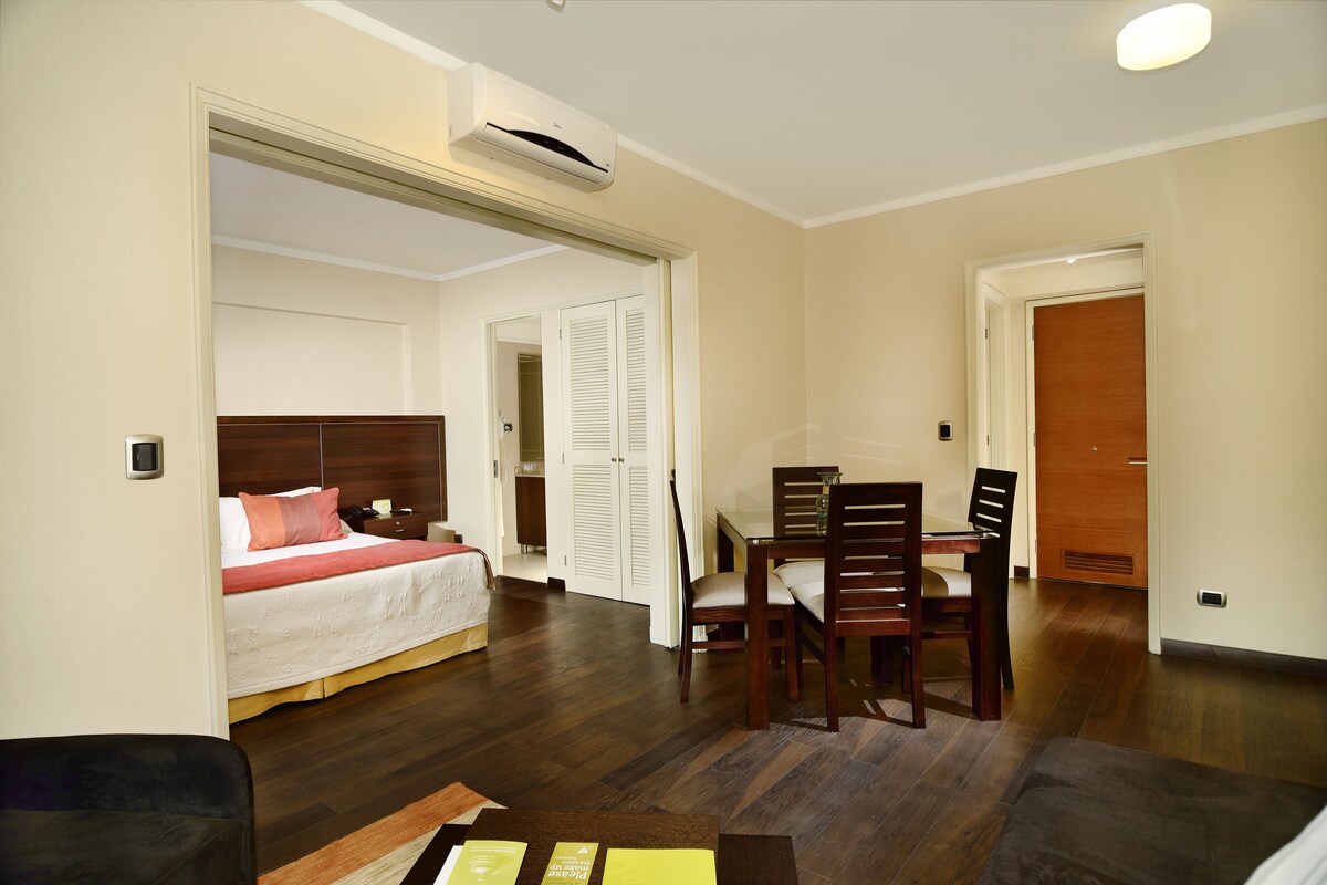 A hotel room with a bed, a dining table with chairs, and a wooden floor.