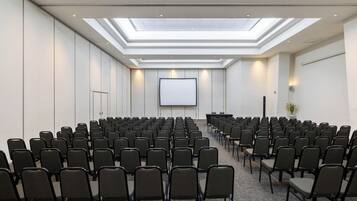 Un salón de conferencias con filas de sillas, una pantalla grande y un tragaluz.
