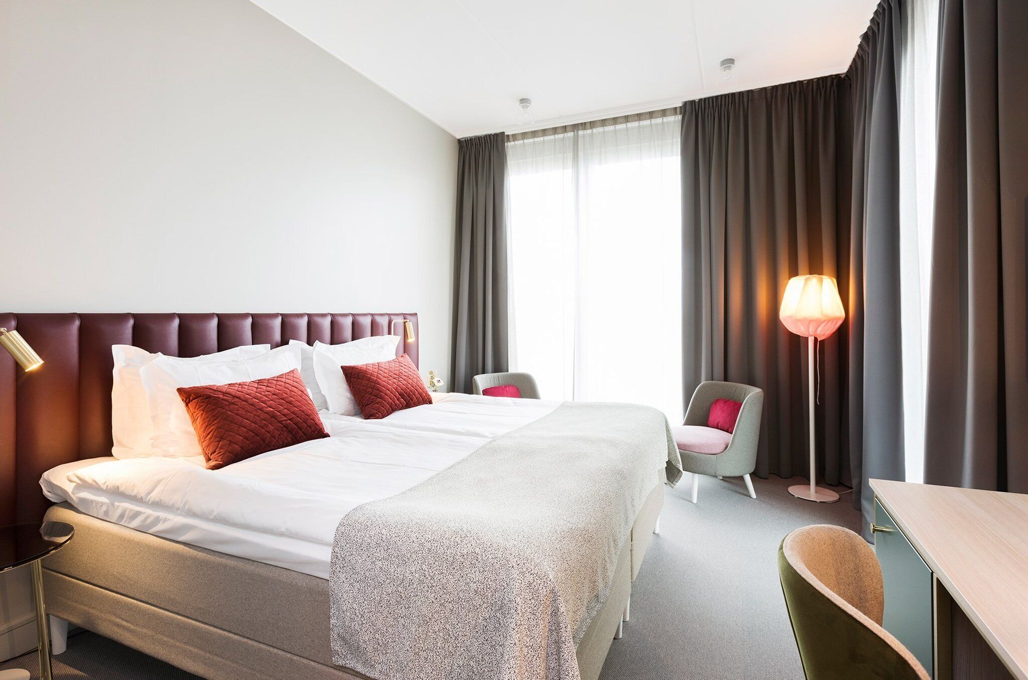 A hotel room with a large bed, two bedside tables, a desk, a chair, and a floor lamp.