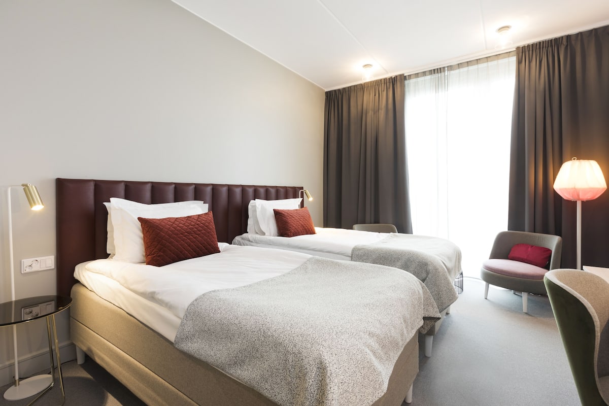 A hotel room with two beds, a desk, a chair, and a lamp.