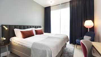 Habitación de hotel con cama, mesita de noche, lámpara, silla y vistas al exterior a través de la ventana.
