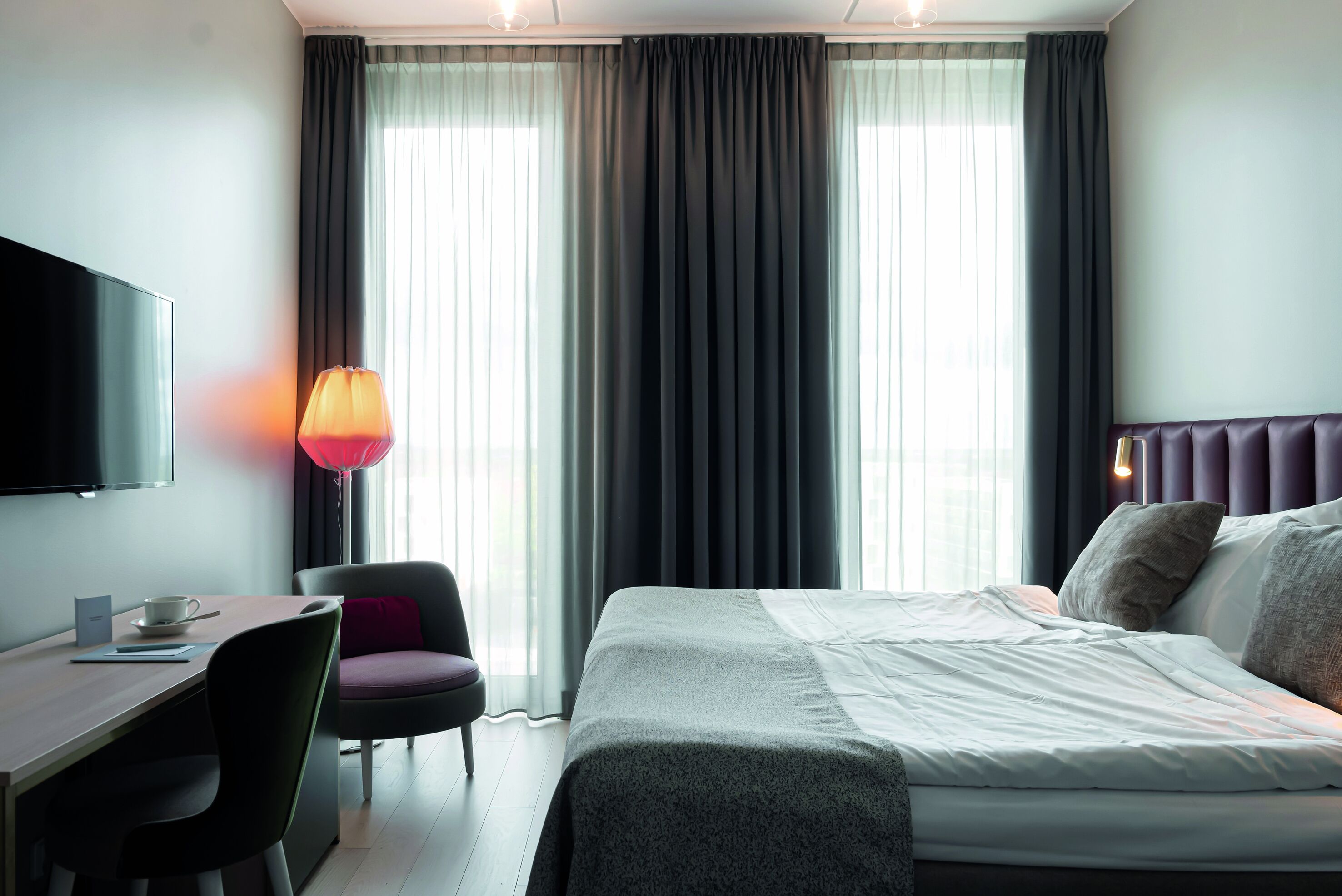 A hotel room with a bed, a desk, a chair, a TV, and a window with curtains.