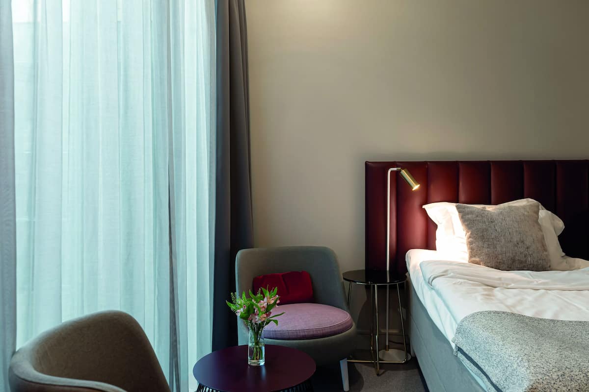 A hotel room with a bed, a chair, a small table with a vase, and a window with curtains.