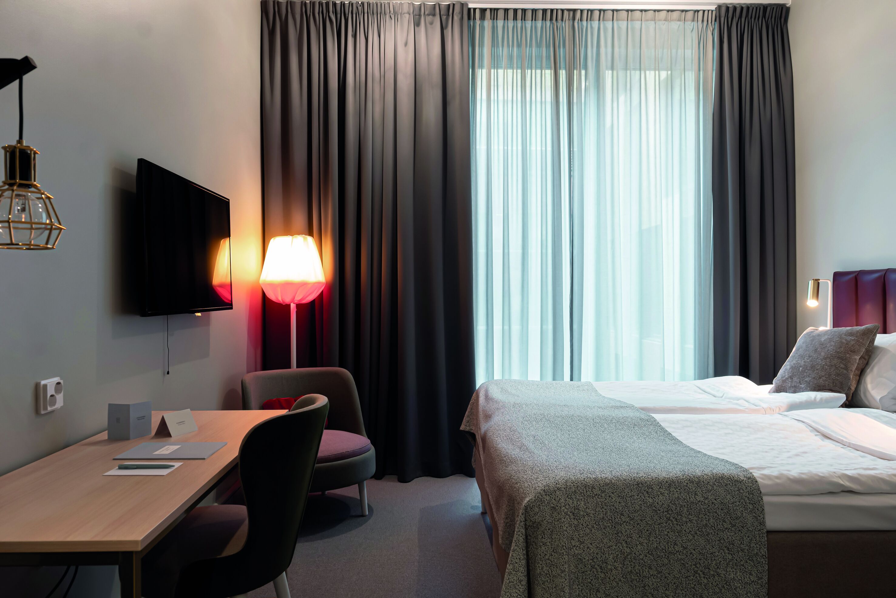 Habitación de hotel con cama, escritorio, silla, televisor y ventana con cortinas.