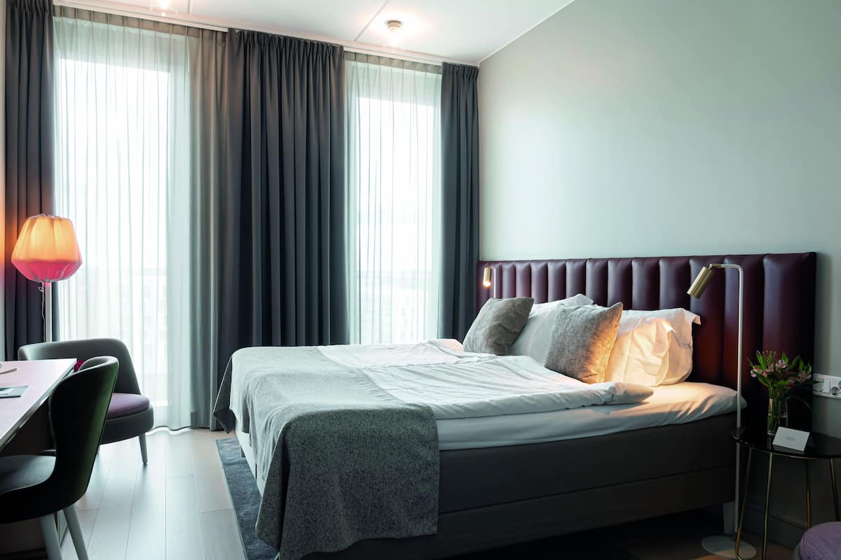 A modern hotel room with a neatly made bed, a desk with a chair, and a bedside table with a lamp.