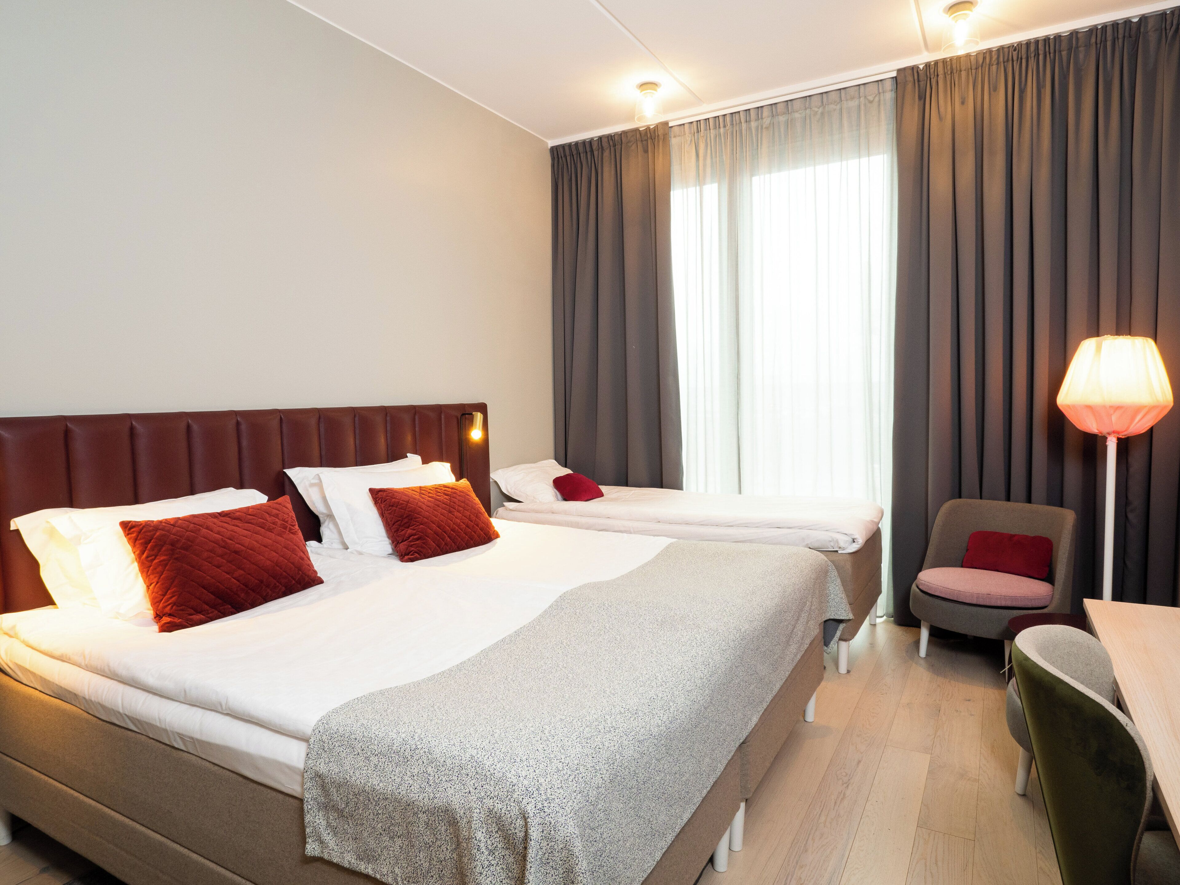 A hotel room with two beds, a desk, a chair, and a lamp.