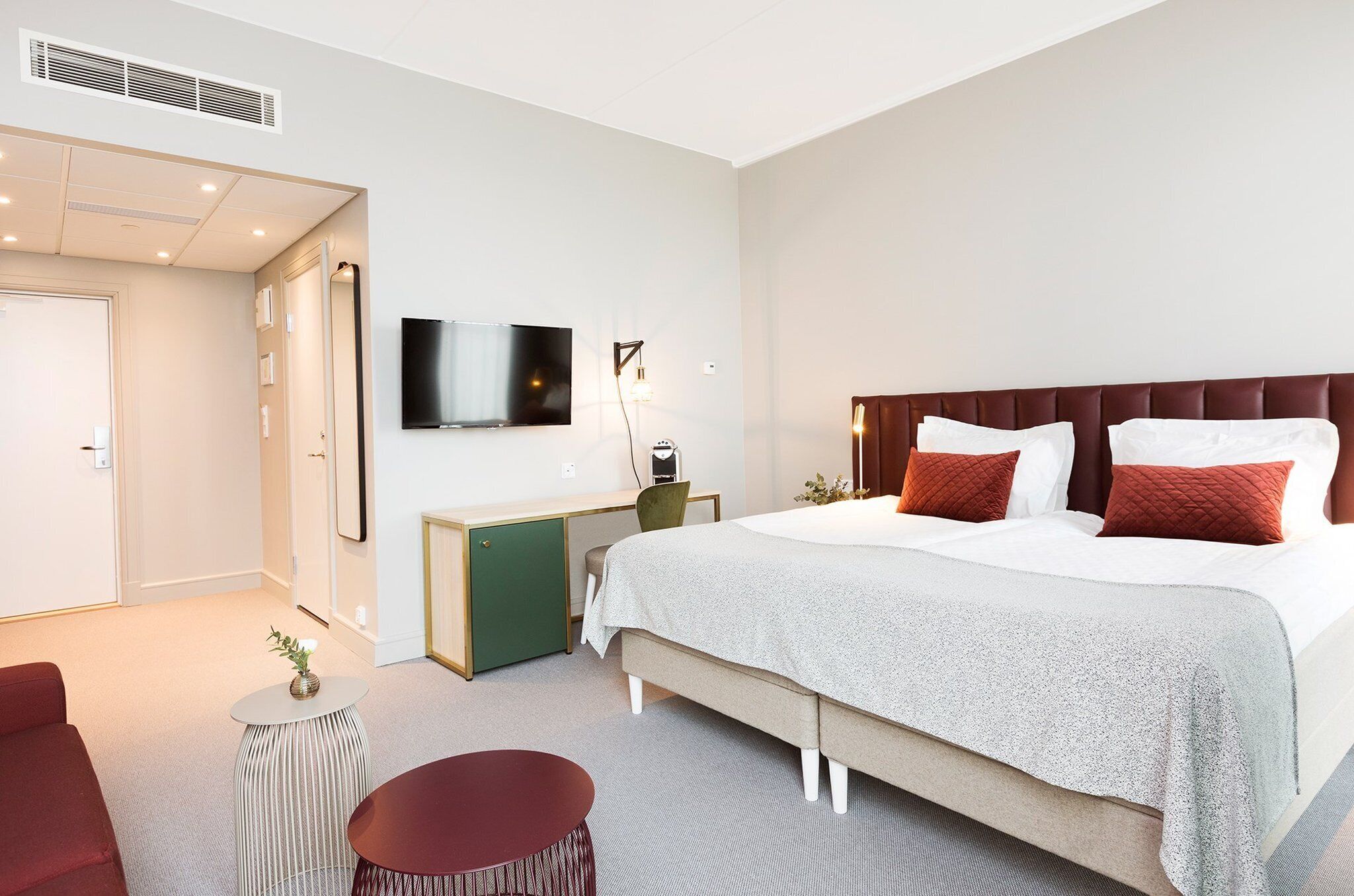 A modern hotel room with a large bed, a desk, a TV, and a small seating area.