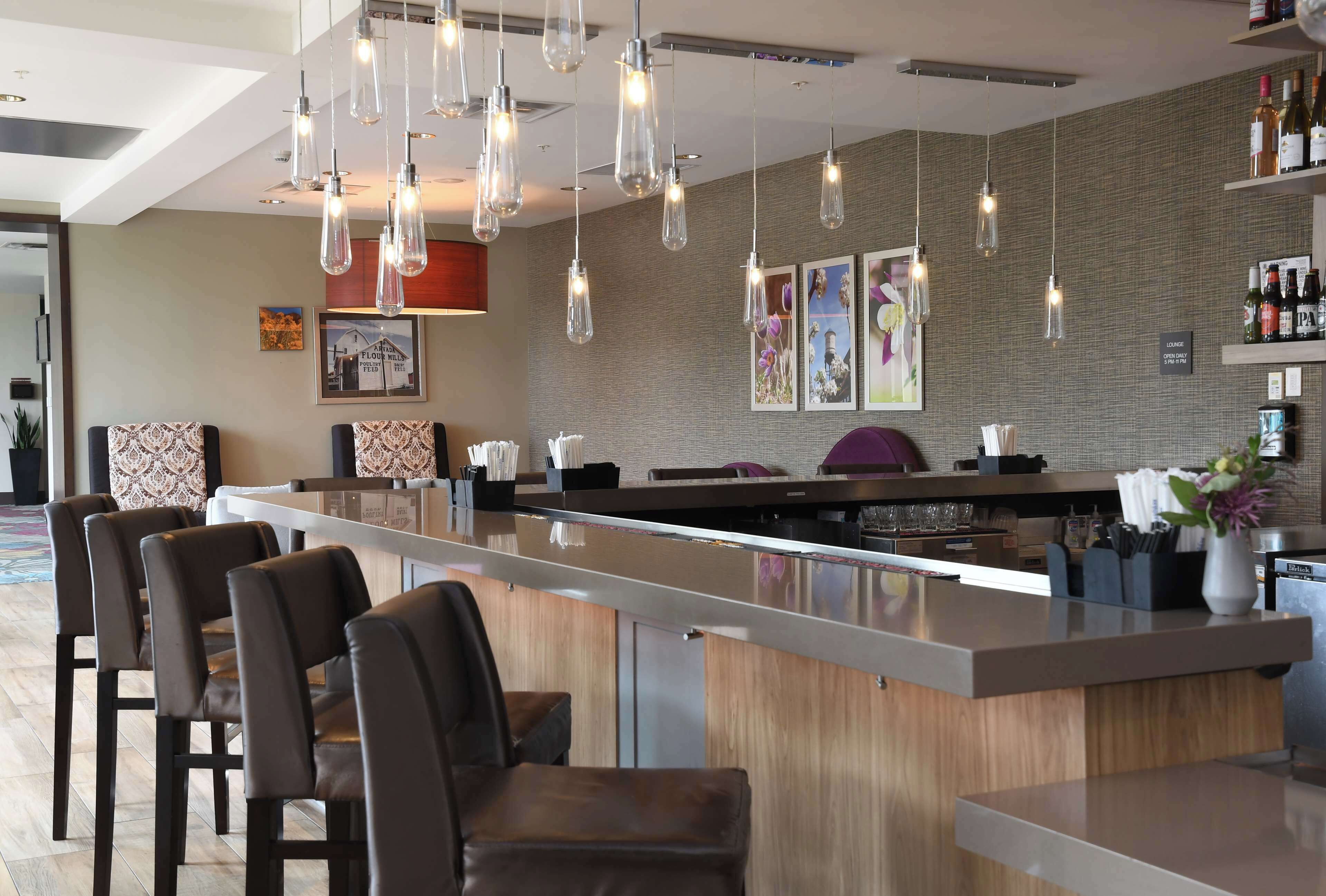 Uma área de bar moderna com um balcão comprido, várias luminárias pendentes e uma parede de tijolos com obras de arte emolduradas.