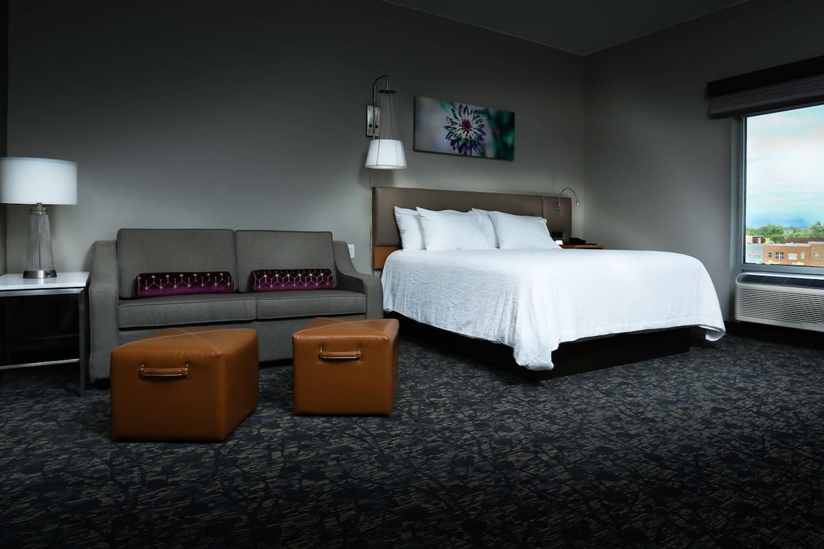 Ein Hotelzimmer mit einem Bett, einem Sofa, einem Sessel, einer Lampe, einem Gemälde und einem großen Fenster.