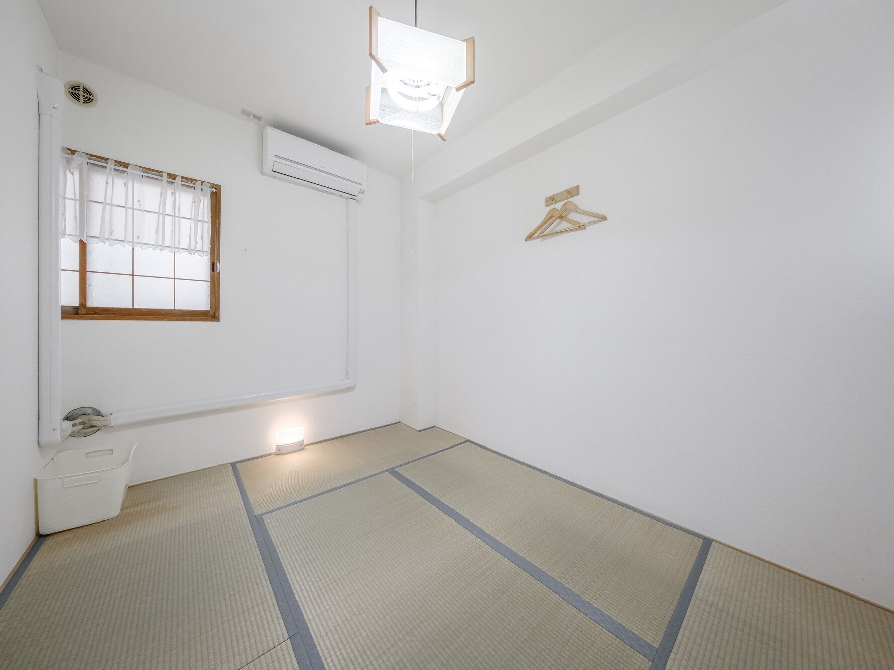 Une pièce avec un sol recouvert de tatamis, une fenêtre avec des rideaux blancs, un climatiseur et un porte-manteau en bois fixé au mur.
