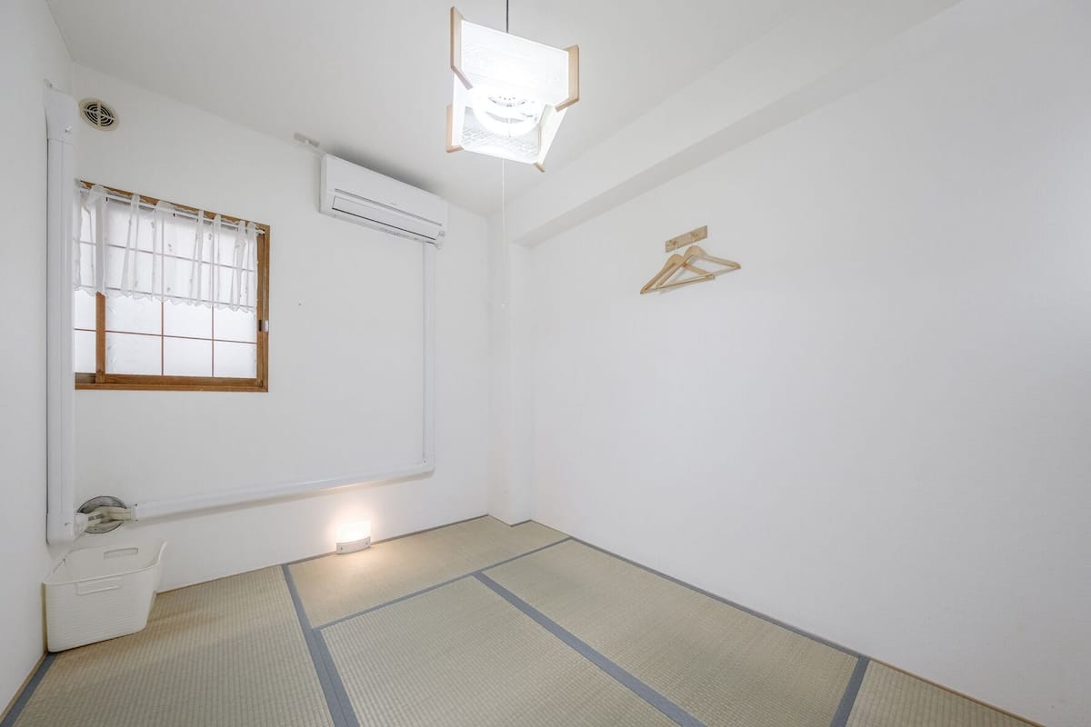 A room with tatami flooring, a window with white curtains, an air conditioning unit, and a wooden hanger on the wall.