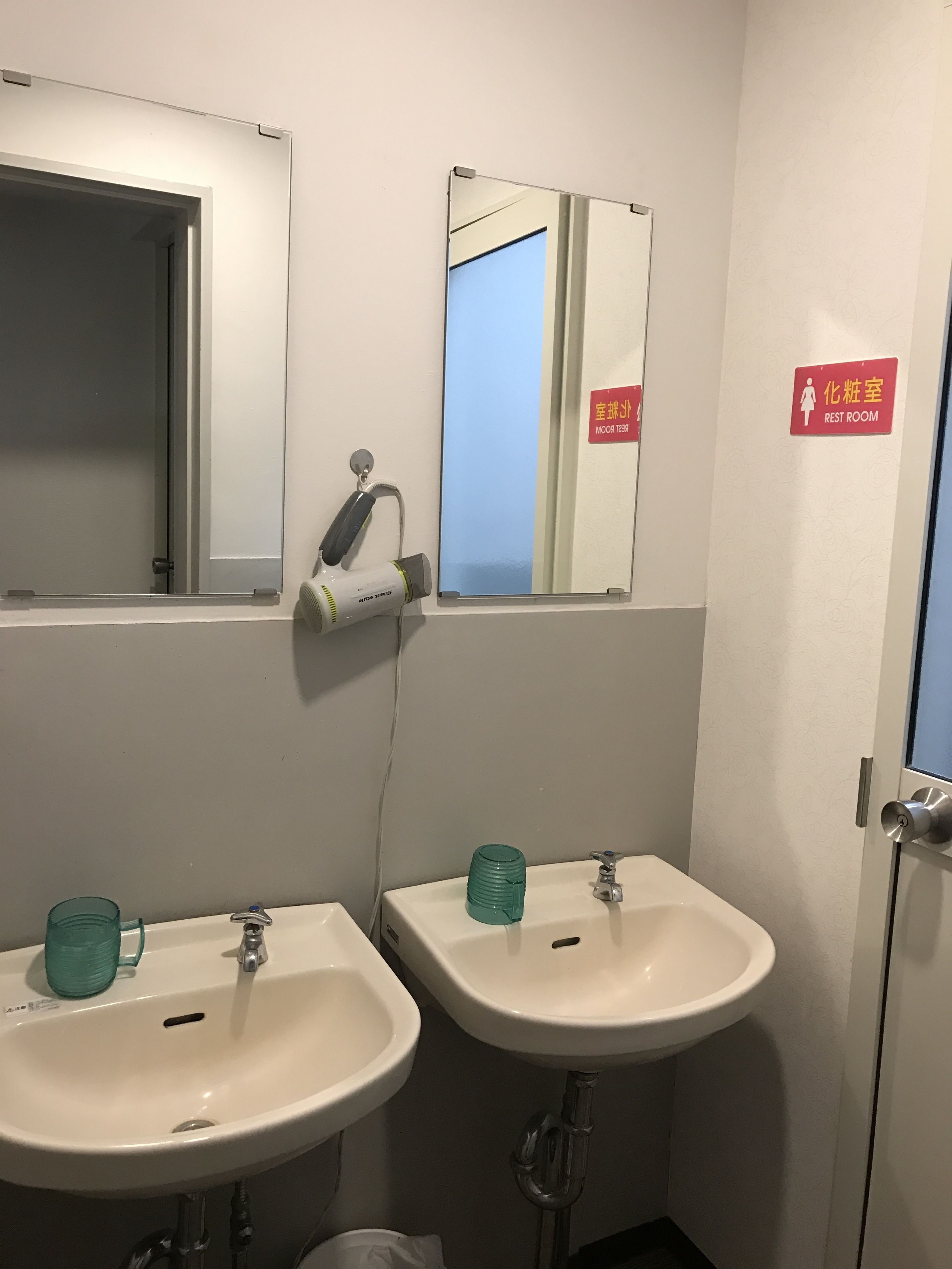 A bathroom with two sinks, a mirror, and a hairdryer. There is a sign indicating a rest room.