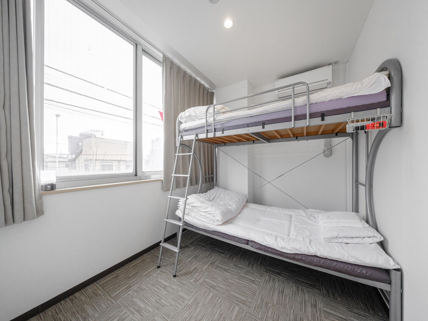 Une chambre avec un lit superposé, une échelle, une fenêtre et une vue sur les bâtiments environnants.