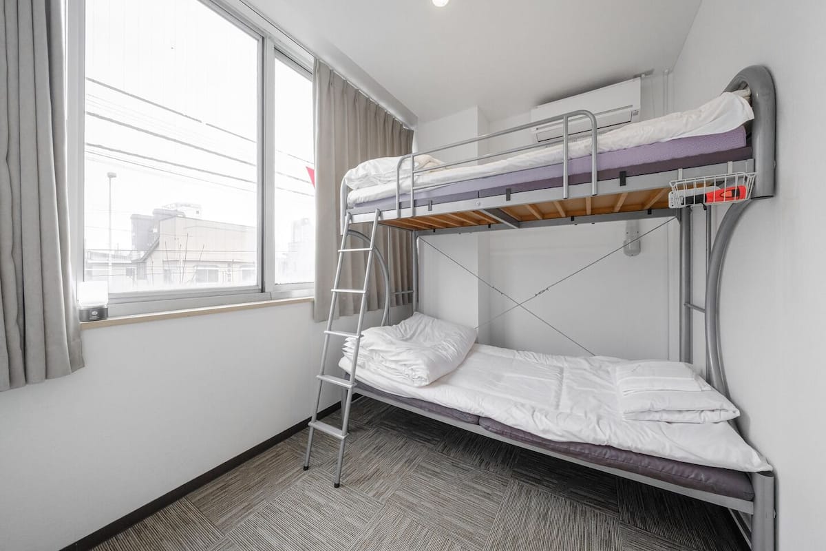 A bunk bed room with a ladder, a window, and a view of buildings outside.