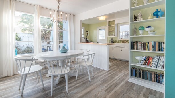 A bright kitchen with white cabinetry, a dining table with chairs, and a bookshelf filled with books.