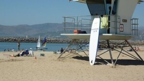 A lifeguard tower with a flag, situated on a sandy beach with a rocky breakwater in the background.