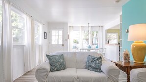 A cozy living room with a white sofa, patterned cushions, a wooden table with a yellow lamp, and a view of the dining area through large windows.