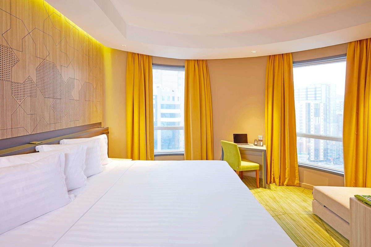 A hotel room with a large bed, a desk with a chair, and a view of the city through large windows.